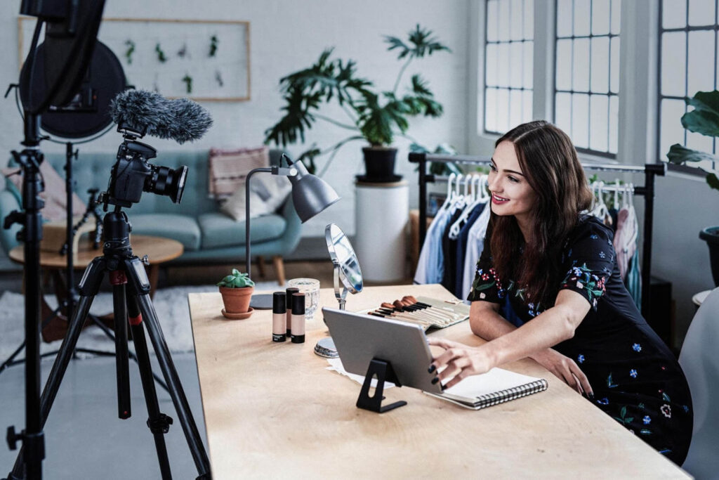 The image shows a young woman sitting at a desk in a home office. She is looking at a tablet and smiling. There is a camera on a tripod in front of her.