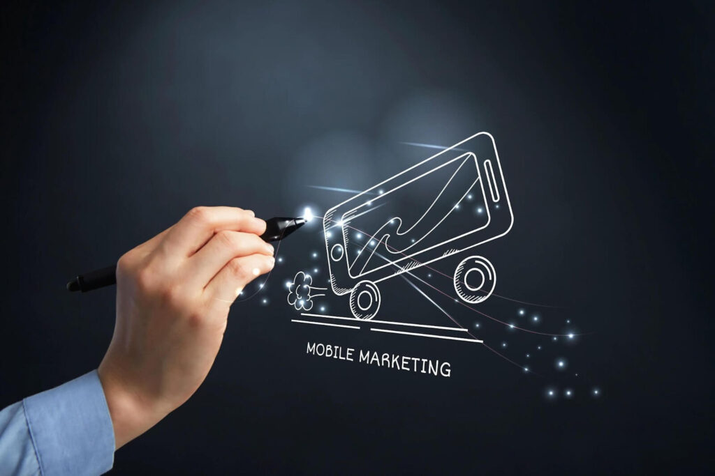 A hand is drawing a smartphone on a blackboard. The smartphone has the words "Mobile Marketing" written on it.