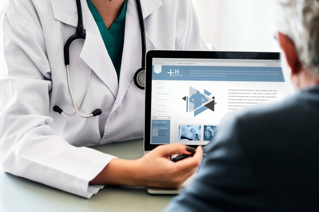 A healthcare professional wearing a white lab coat and stethoscope sits at a desk with a laptop, discussing medical information with a patient seated opposite them.