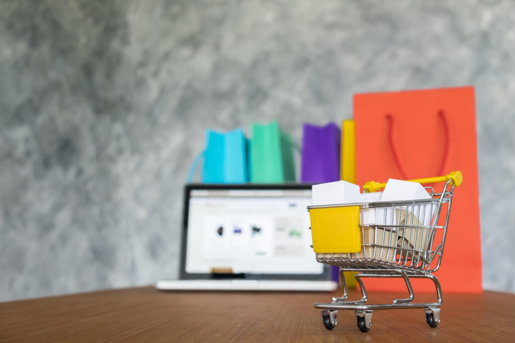 A small shopping cart filled with white boxes sits on a wooden surface. In the background, a laptop displays a shopping website.